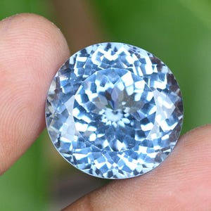 May include: A round, faceted gemstone with a light blue hue. The gem is held between two fingers, showcasing its intricate cut and reflective surfaces. The gem's facets create a sparkling effect, highlighting its clarity and brilliance.
