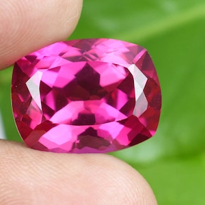 May include: A faceted, cushion-cut gemstone in a vibrant magenta hue. The gem is held between fingers, showcasing its polished surfaces and internal reflections. The background is a soft green, providing contrast to the jewel's color.