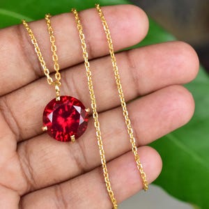 Könnte beinhalten: Eine goldfarbene Halskette mit einem großen, runden, facettierten, tiefroten Edelsteinanhänger. Der Anhänger wird von vier Krappen gehalten. Die Kette hat ein einfaches Kettenglied-Design. Die Halskette wird vor einem verschwommenen Hintergrund gezeigt.