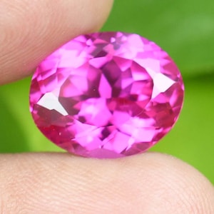 May include: A faceted, round-cut, hot pink gemstone. The gem is held between two fingers against a blurred green background. The gem's facets reflect light, creating a sparkling effect. The gem is a vibrant shade of pink.