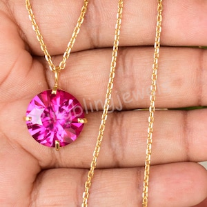 以下が含まれることがあります： 大きな丸いピンク色のファセットペンダントが付いた、ゴールドカラーのチェーンネックレス。ペンダントは4つのゴールドの爪で固定されています。ネックレスはニュートラルな背景に表示されています。