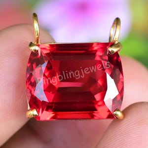 May include: A vibrant red gemstone pendant with a rectangular cut. The gemstone is set in a gold-colored metal frame with small loops at the top for a chain. The text "pingblingjewels" is visible on the gemstone.