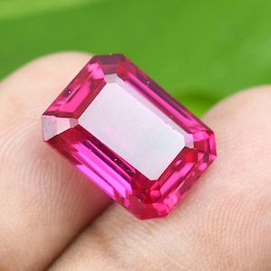 May include: A faceted, rectangular-cut, vibrant pink gemstone. The gem has a bright, reflective surface with a lighter, rectangular interior. The gem is held in a hand against a blurred green background.