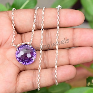 Könnte beinhalten: Eine silberne Halskette mit einem großen, runden, facettierten Amethyst-Anhänger. Der Anhänger hat eine tiefviolette Farbe und hängt an einer feinen Silberkette. Die Halskette wird vor einem verschwommenen grünen Hintergrund gezeigt.