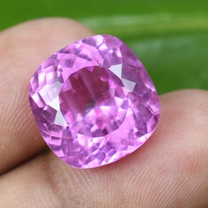 May include: A faceted, cushion-cut, pink gemstone. The gem is held in a hand, with a green background. The gem's facets reflect light, creating a sparkling effect. The gem is a vibrant shade of pink.