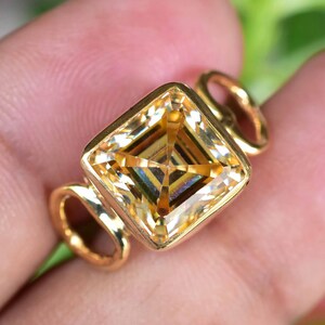 Puede incluir: Un anillo de oro con una gran piedra preciosa amarilla de talla cuadrada. La piedra preciosa está engastada en un engaste de bisel dorado con un diseño geométrico. El anillo tiene un diseño simple y elegante con dos pequeños bucles a cada lado de la piedra preciosa.