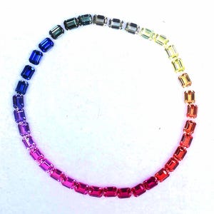 May include: A circular arrangement of rectangular gemstones displaying a spectrum of colours. The gemstones transition from blues and purples to pinks, reds, oranges, yellows, and greens, creating a rainbow effect. Each stone is a different shade.