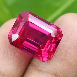 May include: A faceted, rectangular-cut, vibrant pink gemstone. The gem has a high degree of clarity, with multiple facets reflecting light. The gemstone is held in a hand, with a green background.