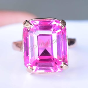 Puede incluir: Un anillo de oro rosa con una gran gema rosa de corte rectangular. La gema está facetada, reflejando la luz, y está engastada con cuatro garras. La banda del anillo tiene un diseño simple y pulido.