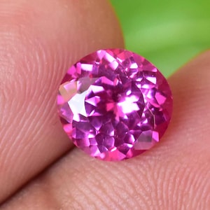 May include: A round, faceted, pink gemstone. The gem is held between fingers, with a green background visible. The gemstone's facets reflect light, creating a sparkling effect. The gem is a vibrant shade of pink.