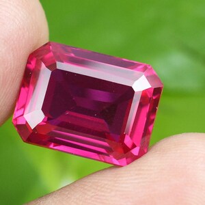 May include: A faceted, rectangular-cut gemstone with a vibrant pink hue. The gem is held between fingers, showcasing its polished surfaces and internal reflections. The gem's color ranges from a deep magenta to a lighter pink.