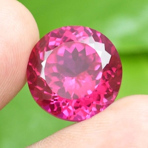 Peut inclure: Une grosse pierre précieuse ronde et facettée d'un rose vif. La gemme est tenue entre deux doigts, mettant en valeur sa taille brillante et ses surfaces réfléchissantes. Les facettes de la pierre précieuse captent la lumière, créant un effet scintillant.