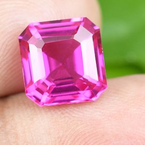 May include: A faceted, rectangular-cut, bright pink gemstone. The gem has a symmetrical design with multiple facets that reflect light. The stone is held in a person's hand, with a green background visible.