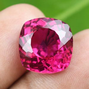 May include: A faceted, cushion-cut gemstone in a vibrant magenta hue. The gem's surface reflects light, showcasing its intricate facets. The gemstone is held against a blurred green background.