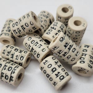 Puede incluir: Cuentas cilíndricas de color marfil con números y líneas negras. Las cuentas están dispuestas en una pila, cada una con un agujero que atraviesa el centro. Los números parecen ser código binario, con una mezcla de 0 y 1.