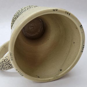 May include: A beige ceramic mug with a wide opening and a rounded handle. The interior is a darker brown. The handle and rim are decorated with small black text. Black markings are visible on the interior of the mug.