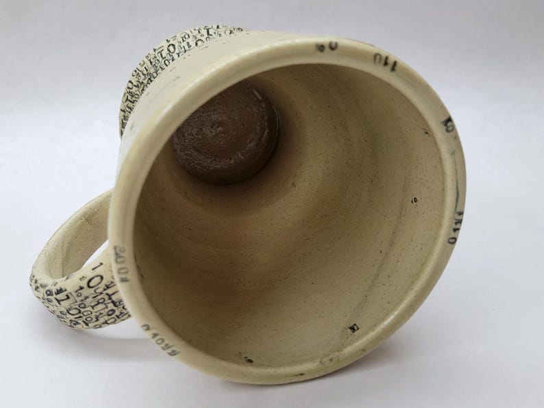 May include: A beige ceramic mug with a wide opening and a rounded handle. The interior is a darker brown. The handle and rim are decorated with small black text. Black markings are visible on the interior of the mug.