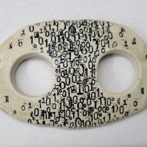 May include: An oval-shaped off-white ceramic toggle button featuring two circular openings. The surface is decorated with a dense, black printed pattern of binary code consisting of numerous zeros and ones scattered across the front face.