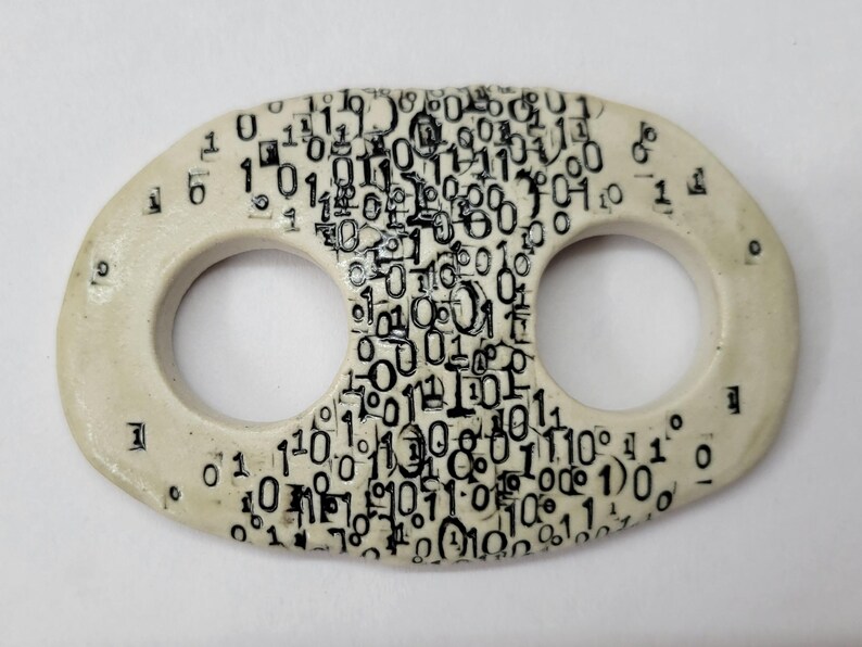 May include: An oval-shaped off-white ceramic toggle button featuring two circular openings. The surface is decorated with a dense, black printed pattern of binary code consisting of numerous zeros and ones scattered across the front face.