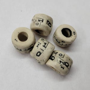 May include: Four off-white cylindrical beads with a matte finish, each featuring black printed numbers and symbols including ones, zeros, and sixes. The beads have a central hole and a slightly textured surface, arranged in a small cluster.