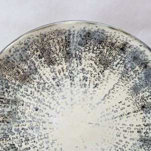May include: A ceramic plate with a white centre and a dark blue and grey rim. The plate is decorated with a pattern of small black numbers and symbols radiating from the centre. The rim has a silver metallic finish.