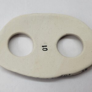 May include: An off-white, oval-shaped ceramic button featuring two circular holes and the number 01 printed in black ink between the holes. The surface has a smooth, matte finish with subtle speckling throughout the material.