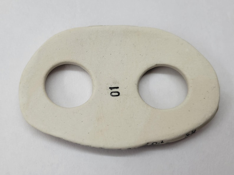 May include: An off-white, oval-shaped ceramic button featuring two circular holes and the number 01 printed in black ink between the holes. The surface has a smooth, matte finish with subtle speckling throughout the material.