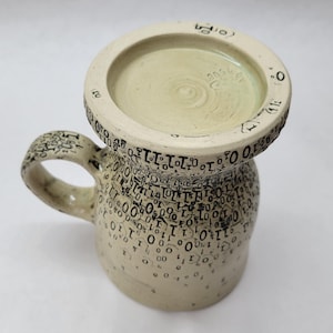 May include: A handmade ceramic mug with a wide base and a circular top. The mug is off-white with black numbers and symbols covering the surface. The handle and rim also feature the same black detailing. The interior is a light green colour.