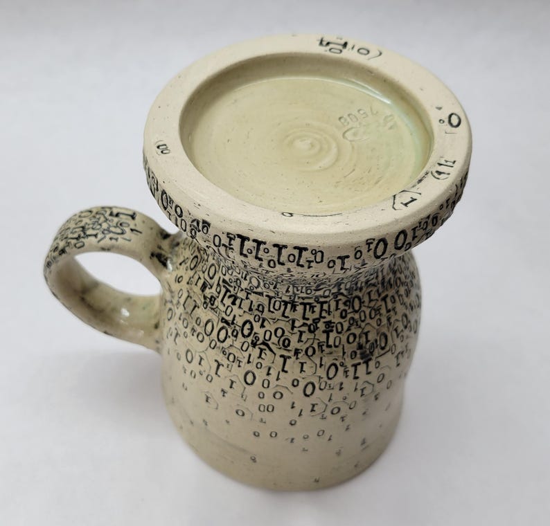 May include: A handmade ceramic mug with a wide base and a circular top. The mug is off-white with black numbers and symbols covering the surface. The handle and rim also feature the same black detailing. The interior is a light green colour.