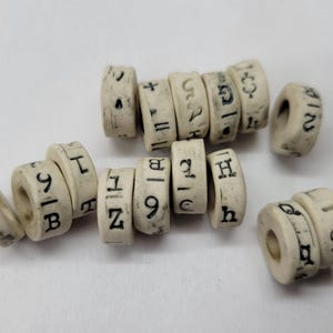 May include: Ivory-colored, cylindrical beads with black markings, including numbers, letters, and symbols. The beads are arranged on a white surface. The markings appear to be hand-painted or stamped. The beads are likely used for crafts or jewelry making.
