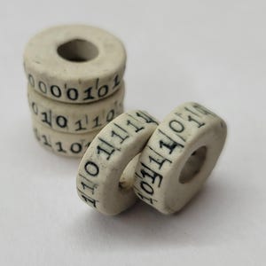 May include: A stack of off-white, donut-shaped beads with black binary code printed around the edges. The beads are arranged in a stack of four, with two additional beads lying separately.