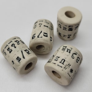 May include: Four off-white cylindrical beads with black symbols and letters. Each bead has a hole running through the centre. The beads are arranged on a white surface.