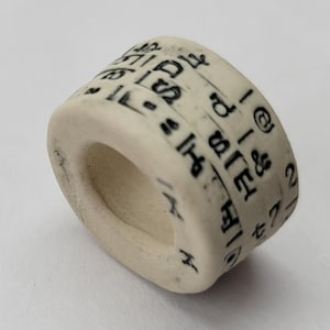 May include: A cream-colored, cylindrical ring with black symbols and numbers etched around its exterior. The ring has a hollow center and appears to be made of ceramic or a similar material. The symbols are small and evenly spaced.