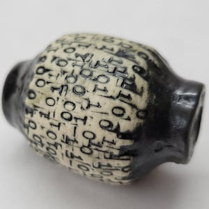 May include: A ceramic, barrel-shaped decorative object. The central body is off-white with a repeating pattern of black symbols. The ends are solid black. The object has a slightly rough texture.