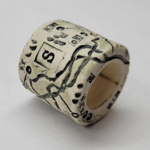 May include: A cylindrical, off-white ceramic ring with a textured surface. The ring features black, stylized map-like designs and the letter "S" in a square. The ring is likely handmade and has a rustic aesthetic.