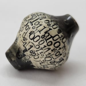 May include: A small, rounded ceramic bead featuring a cream-colored surface decorated with a repeating black binary code pattern of ones and zeros. The bead has a smooth, glossy finish with dark, charcoal-colored accents at both ends of the piece.