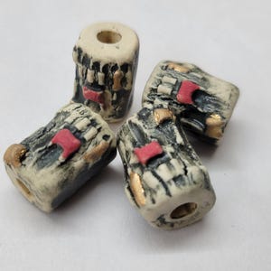 May include: Four ceramic beads with a cylindrical shape. The beads have a cream-colored base with black and gold accents. Each bead features a small, rectangular red detail. The beads are designed with a textured, rustic appearance.