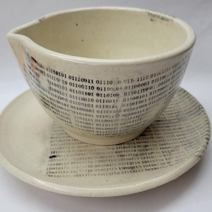 May include: A cream-colored ceramic bowl with a pouring spout and a matching saucer. Both pieces feature a repeating black print of binary code consisting of zeros and ones. The ceramic has a speckled texture throughout the surface of the bowl and saucer.