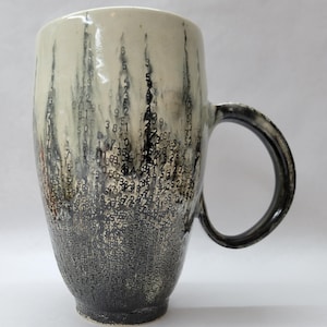 May include: A tall ceramic mug with a unique design. The mug features a cream-colored top that transitions to a dark gray base. The surface is covered with a pattern of black numbers. The handle is dark gray. A decorative and functional item.