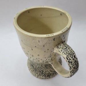 May include: A handmade ceramic mug with a beige glaze. The mug is decorated with black numbers and symbols, including binary code on the handle and base. The rim of the mug has numbers and symbols. The mug has a unique, artistic design.