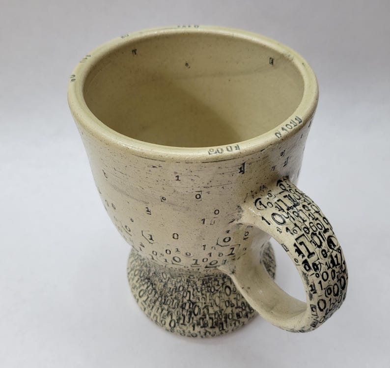 May include: A handmade ceramic mug with a beige glaze. The mug is decorated with black numbers and symbols, including binary code on the handle and base. The rim of the mug has numbers and symbols. The mug has a unique, artistic design.