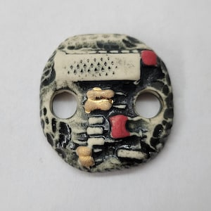 Peut inclure: Perle en céramique de forme ovale avec deux trous. La perle présente une surface texturée avec des accents noirs, blancs, rouges et dorés. Le motif comprend des formes et des motifs géométriques, avec un aspect unique et artisanal.