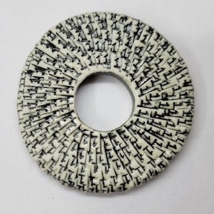 May include: A circular pendant featuring a cream-colored surface with a radial pattern of black, incised, abstract markings. The object has a smooth, rounded outer edge and a central circular cutout, creating a ring-like shape with a textured finish.