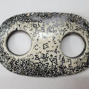 May include: An oval-shaped ceramic toggle button with a glossy off-white finish. The surface is densely covered in a black printed pattern of various numbers and mathematical symbols. The button features two circular holes positioned in the center.