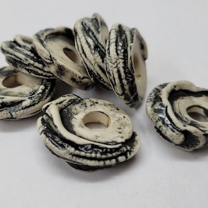 May include: A collection of six off-white, ring-shaped beads with dark gray, organic-looking patterns. Each bead has a central hole. The beads are arranged on a white surface, suggesting a craft or jewelry supply.