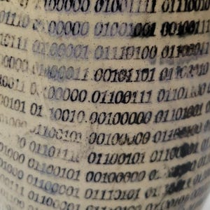 May include: A cylindrical object with a cream-colored surface featuring a repeating pattern of black binary code digits. The text consists of various sequences of zeros and ones printed in a dark, slightly weathered font across the curved surface.