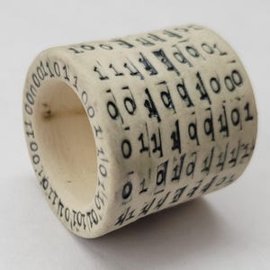May include: A cylindrical object with a cream-colored surface, inscribed with black binary code and other symbols. The object has a hollow center and appears to be made of a solid material. The text is evenly spaced around the cylinder.