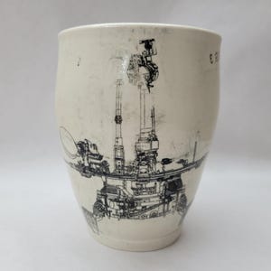 May include: Off-white ceramic tumbler with a black illustration of a complex machine, possibly a spacecraft or industrial equipment. The design wraps around the cup, with details including antennas and mechanical components. The cup has a slightly textured surface.