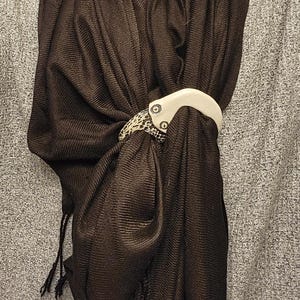 May include: A dark brown ribbed scarf secured with a two-toned scarf ring. The ring features a smooth, off-white curved top section attached to a textured, black and white speckled lower section by two visible silver-toned metal screws.
