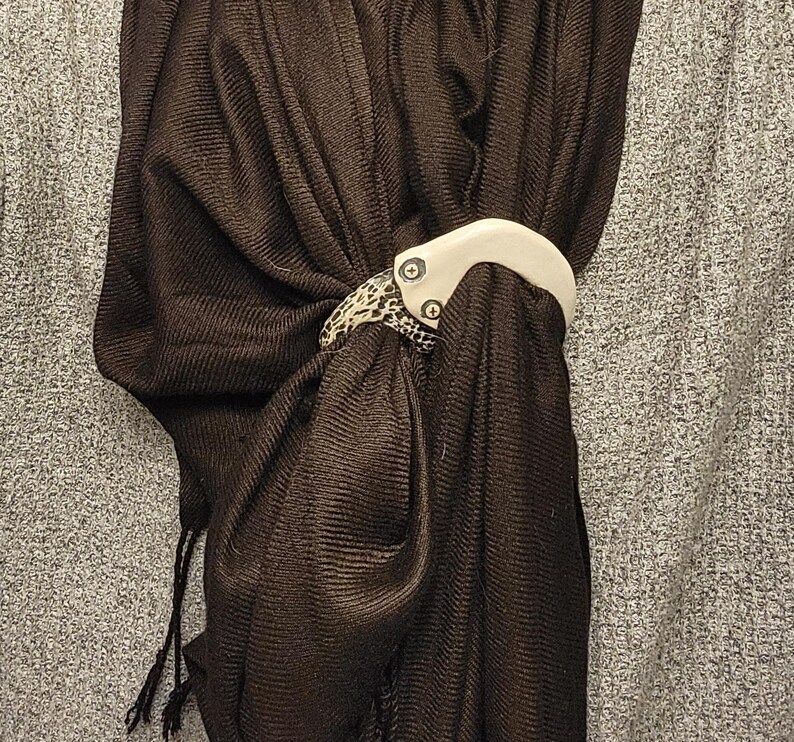 May include: A dark brown ribbed scarf secured with a two-toned scarf ring. The ring features a smooth, off-white curved top section attached to a textured, black and white speckled lower section by two visible silver-toned metal screws.
